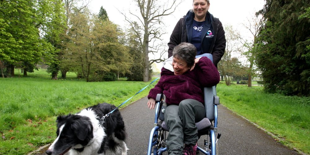 A live-in carer taking an elderly woman with disabilities in a wheelchair to a park with a dog