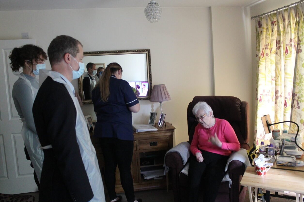 An elderly person in their home sitting in an armchair supported by three care providers