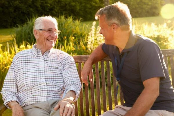 An elderly man and Abbots Care worker sitting on a bench in a park smiling